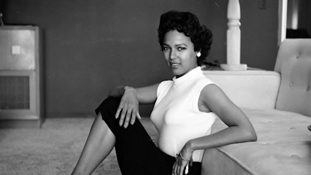 Black and white photo of a young Dorothy Dandridge posing elegantly on the floor, wearing a fitted white top and black pants