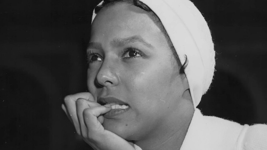 Black and white photo of Dorothy Dandridge, gazing thoughtfully to the side while biting her fingernail, with a scarf wrapped around her head