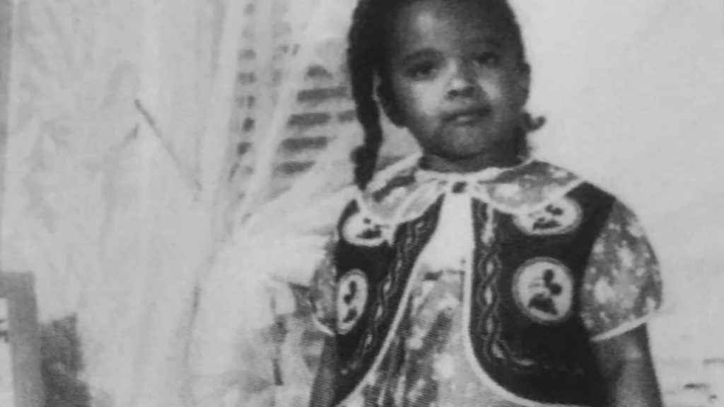 Black and white photo of young Harolyn Suzanne Nicholas wearing a decorative dress, looking seriously at the camera