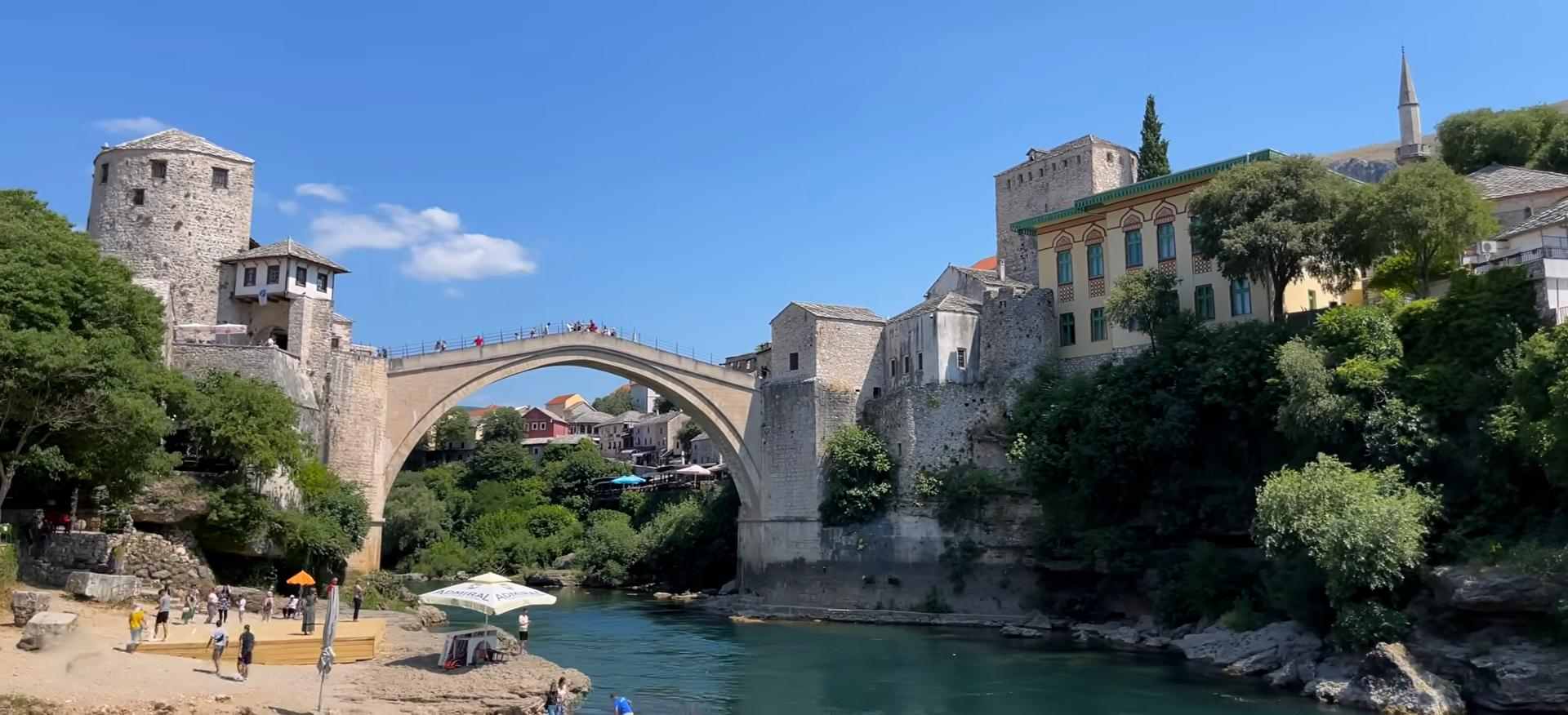 Stone arch bridge over a river in Mostar, surrounded by historic buildings and greenery.